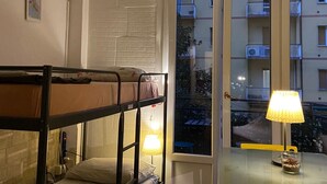 A dorm-style room with bunk beds, a table with a lamp, and a view of a balcony with other apartments. - Europe station rooms (Parma)