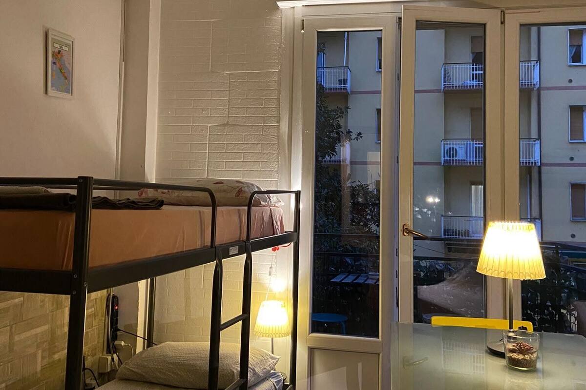 A dorm-style room with bunk beds, a table with a lamp, and a view of a balcony with other apartments.
