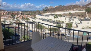 A balcony with a table and chairs, overlooking a parking area and a town with buildings and hills.