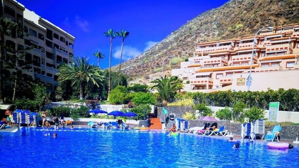 A swimming pool with lounge chairs and a water slide, surrounded by buildings and palm trees.