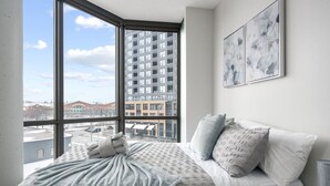 A bedroom with a large window, a bed with patterned bedding, and two abstract wall art pieces.