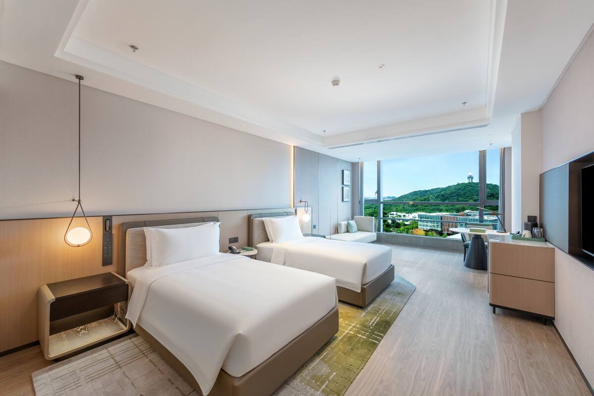 Ein modernes Hotelzimmer mit zwei Betten, einem Schreibtisch, einem Fernseher und einem Balkon mit Blick auf die Stadt.