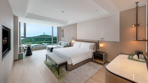 A modern hotel room with a large bed, a sitting area with a sofa and chairs, a small table, and a view of a hill through large windows. - Nanjing Yangzi River Conference Center Longshan Lake Hotel (Nanjing)