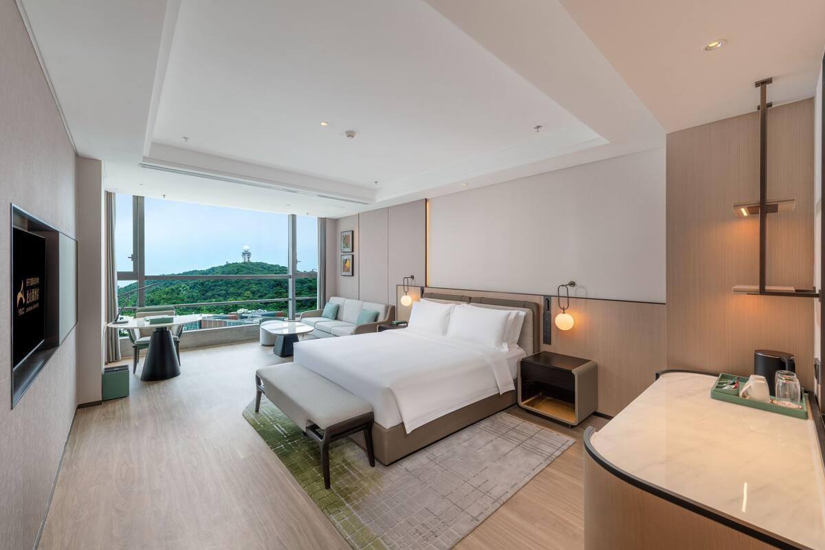 Ein modernes Hotelzimmer mit einem großen Bett, einer Sitzecke mit Sofa und Sesseln, einem kleinen Tisch und Blick auf eine Hügel landschaft durch große Fenster.