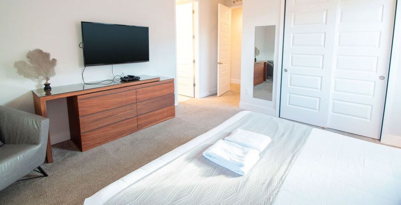 A bedroom with a bed, a TV mounted on the wall, a wooden dresser, and a door.