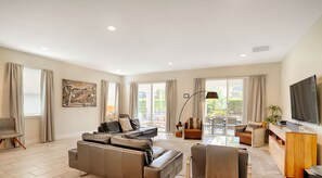 A spacious living room with a sofa, armchairs, a TV, and a balcony with outdoor furniture.