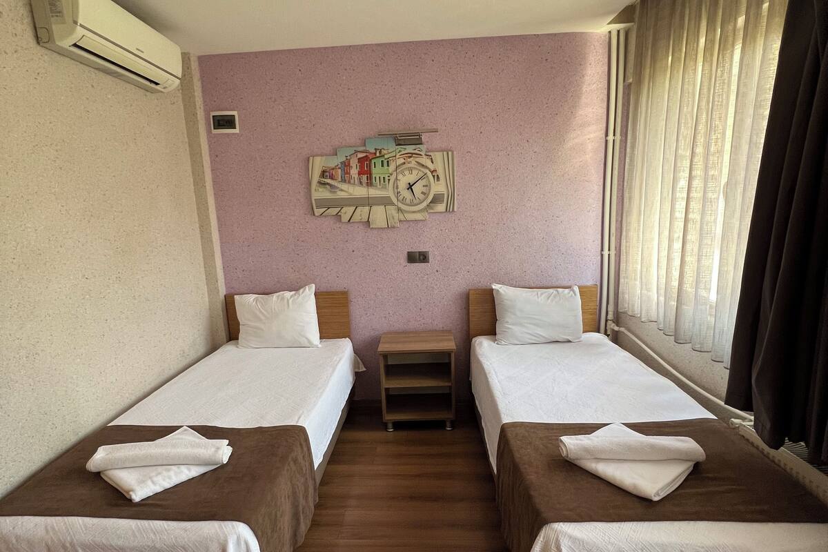 A hotel room with two single beds, a small wooden nightstand, a wall clock, and a window with curtains.