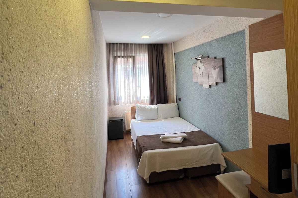 A hotel room with a bed, a small desk, a radiator, and a window with curtains.