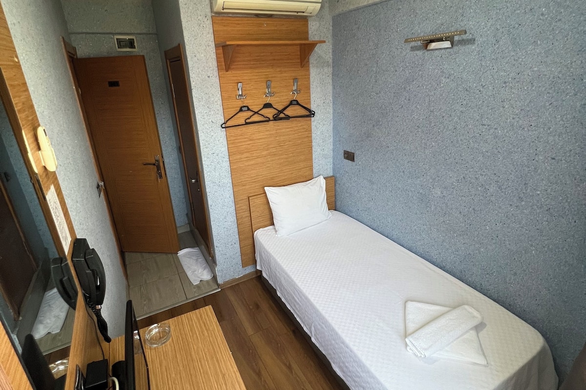 A compact hotel room with a single bed, a wooden desk, a mirror, and a wall-mounted air conditioner.