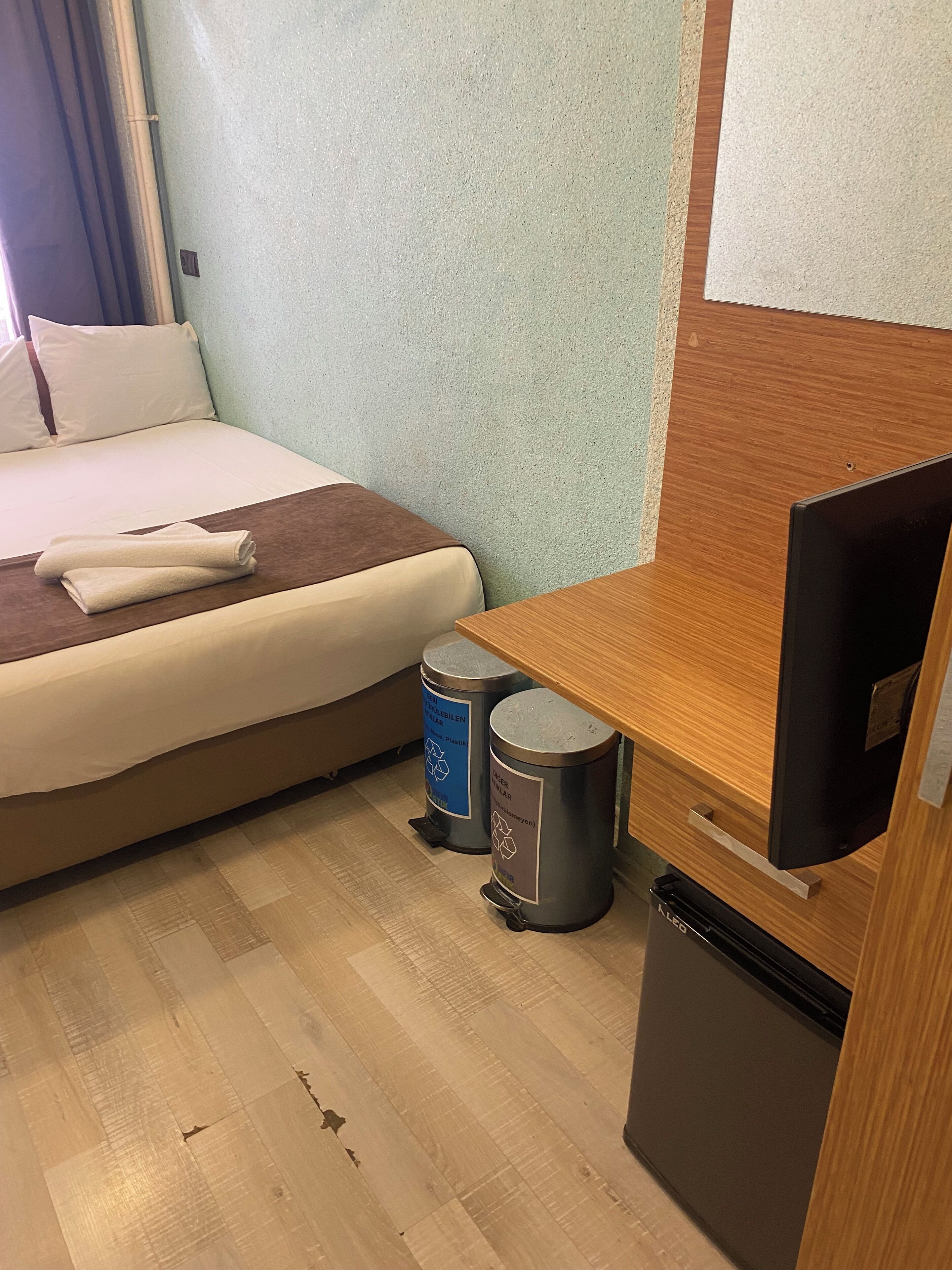 A hotel room with a bed, a wooden desk, a television, and two trash bins.