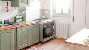 Une cuisine équipée d’une cuisinière, d’un four et de meubles de rangement verts. On y trouve une fenêtre à cadre blanc et un luminaire suspendu.