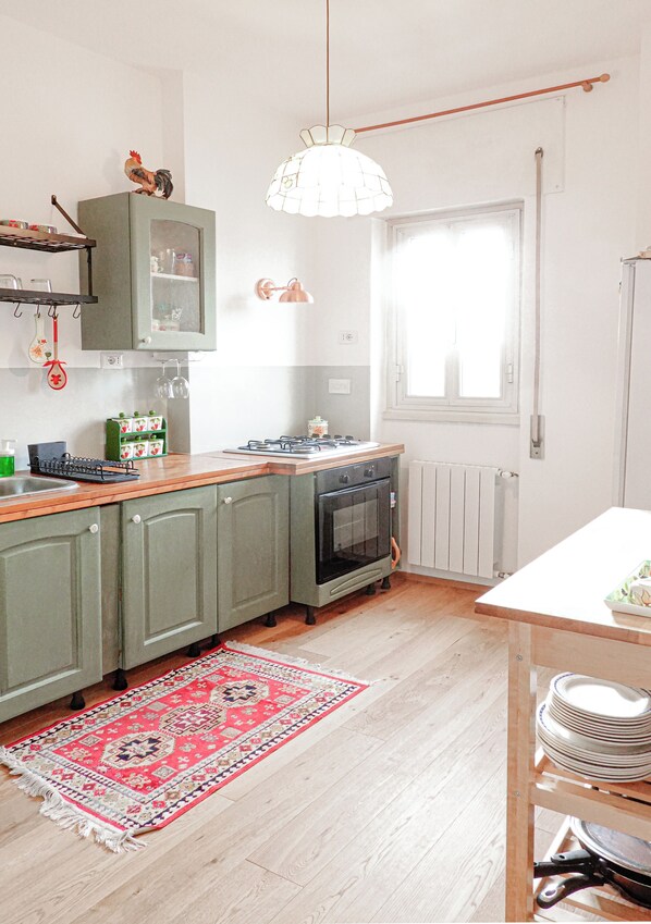 Eine Küche mit Herd, Backofen und grünen Schränken. Ein Fenster mit weißem Rahmen und eine Pendelleuchte sind vorhanden.