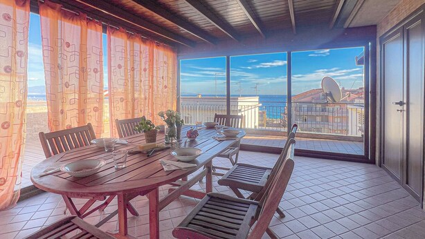 A dining area with a wooden table and chairs, a view of the sea through large windows, and a balcony with a view of the cityscape. - Beach House Roccalumera (Roccalumera)