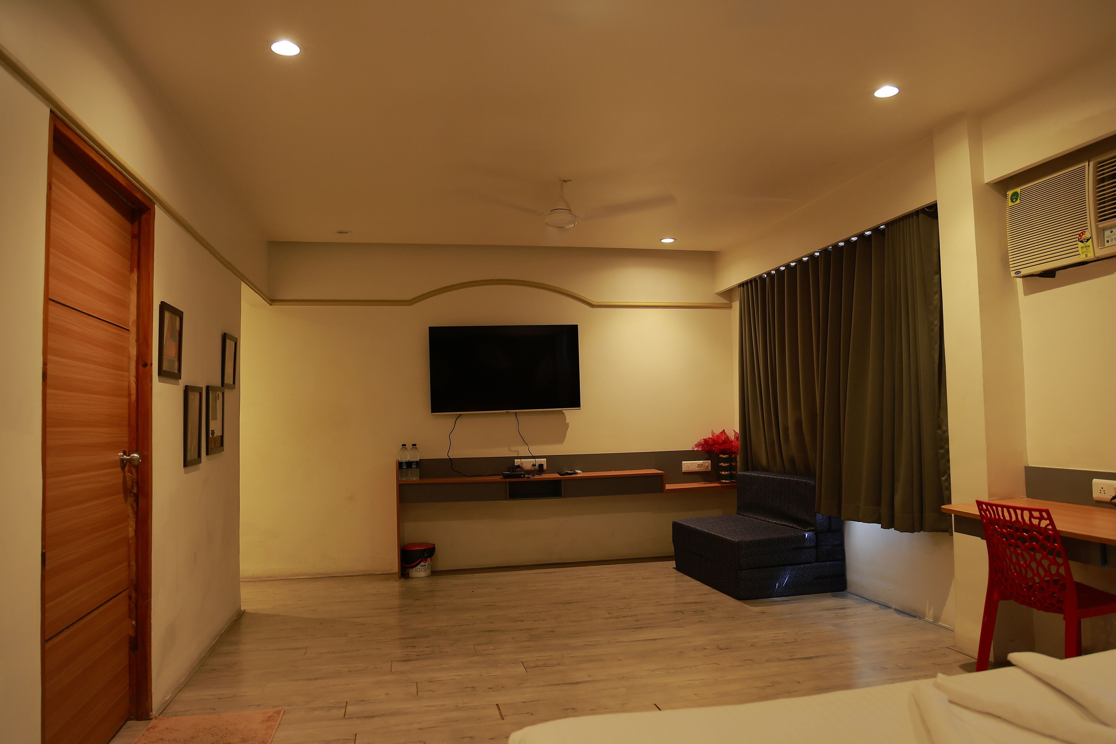 A hotel room with a wooden floor, a flat-screen TV mounted on the wall, a desk with a chair, a sofa, and a window with curtains.
