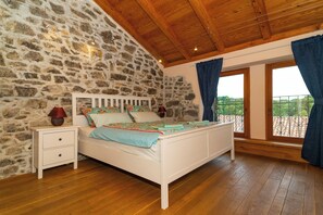A bedroom with a stone wall, wooden ceiling, a white bed, and a view through a window with curtains. - Luxurious Villa in Ružići Croatia with Private Pool and Garden (Ružici)