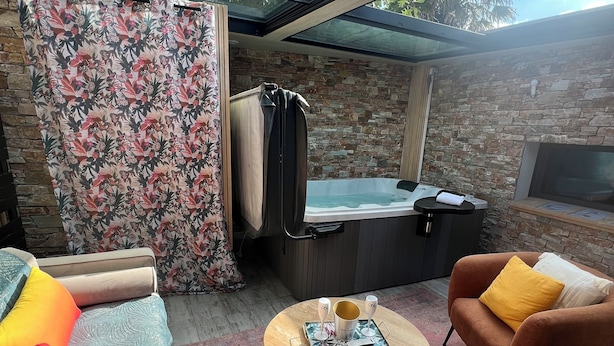 A modern outdoor living area with a glass-enclosed space, a hot tub, a fireplace, and a sofa with colorful cushions. - Love Room with private spa  (Le Perrier)