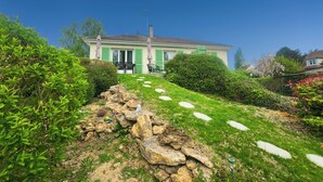 Ein Haus mit grüner Fassade, ein Steinweg, der zu einer Terrasse mit Gartenmöbeln führt, und ein klarer, blauer Himmel.