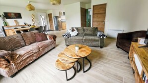 Ein geräumiges Wohnzimmer mit einer großen Couch, einer kleineren Couch, einem runden Couchtisch und einem dunklen Sessel. Im Hintergrund befindet sich ein Essbereich mit einem Holztisch und Stühlen.