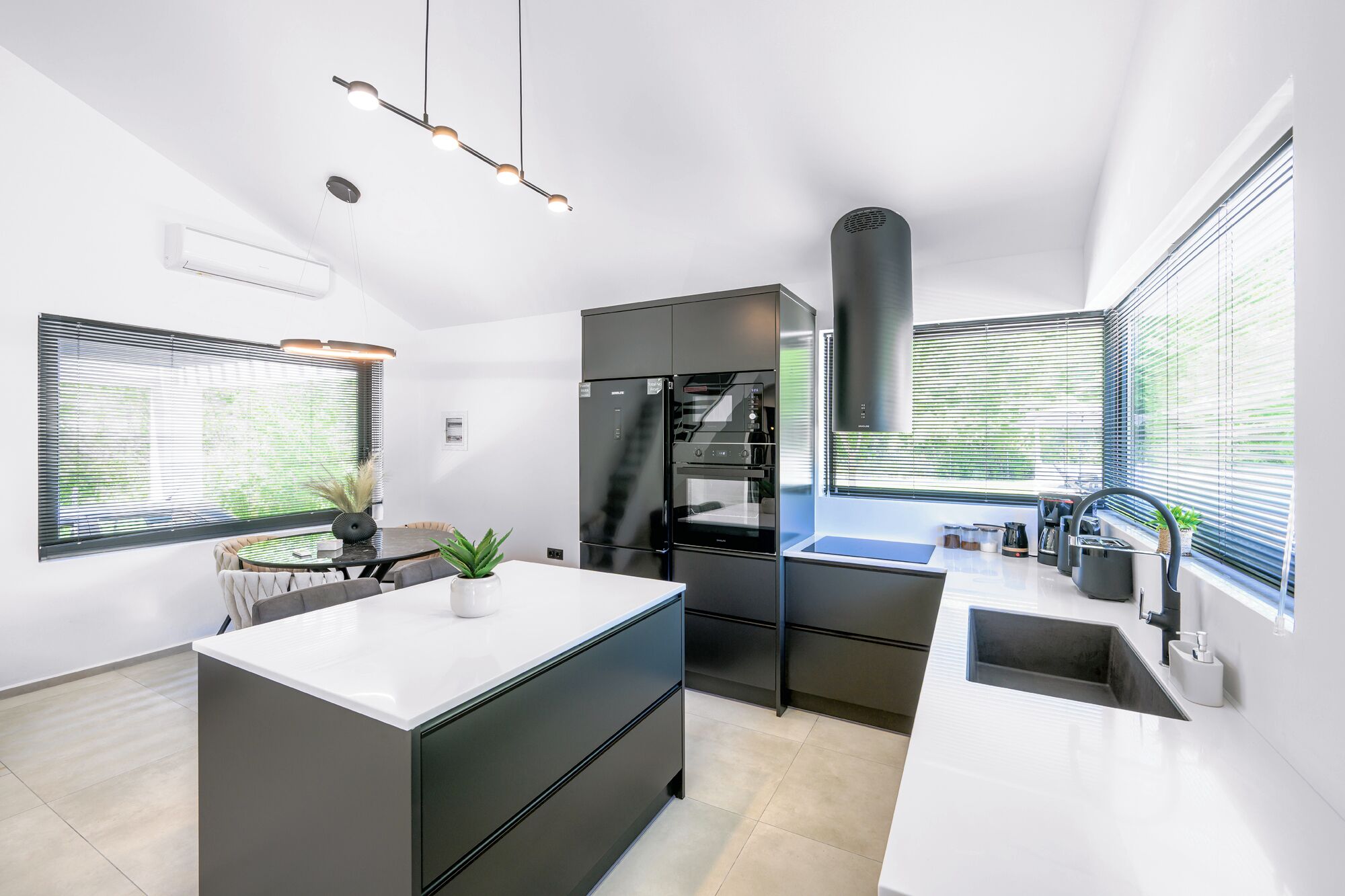 Une cuisine moderne dotée d’un îlot central, de meubles de rangement noirs et d’un four encastré. On y trouve des fenêtres équipées de stores, un coin repas avec une table et des chaises, ainsi qu’un luminaire suspendu.
