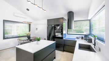 Une cuisine moderne dotée d’un îlot central, de meubles de rangement noirs et d’un four encastré. On y trouve des fenêtres équipées de stores, un coin repas avec une table et des chaises, ainsi qu’un luminaire suspendu.