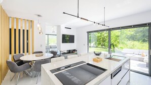 Een moderne keuken met een centraal eiland, een eetgedeelte met grijze stoelen en een groot raam met uitzicht op groen.