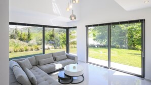 Een moderne woonkamer met een hoekbank, een ronde glazen salontafel en grote ramen met uitzicht op een weelderige tuin en heuvels.