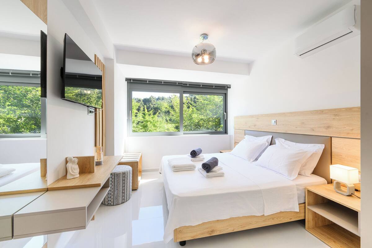 A modern bedroom with a bed, bedside tables, a TV, and a window with a view of greenery.
