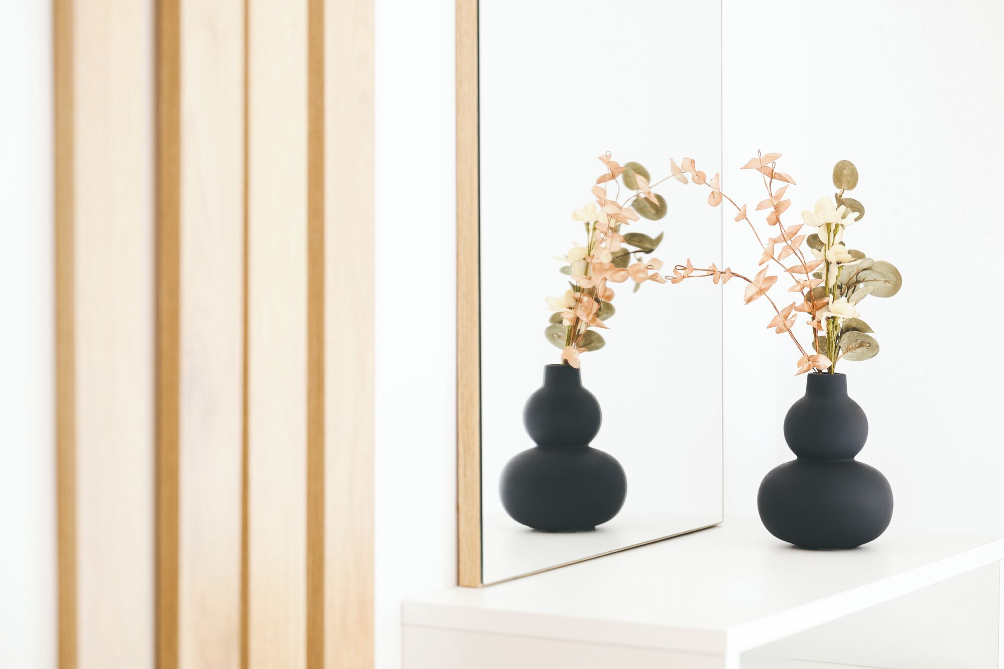 Dois vasos pretos com arranjos florais em uma prateleira branca, refletidos num espelho.
