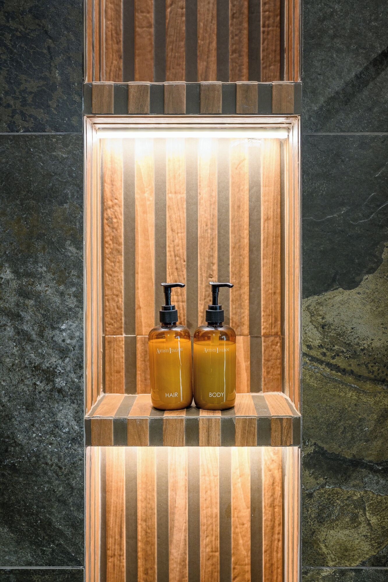 Dos botellas color ámbar, etiquetadas 'Hair' y 'Body', con dos dosificadores negros, sobre una superficie reflectante y un fondo de panel de madera.