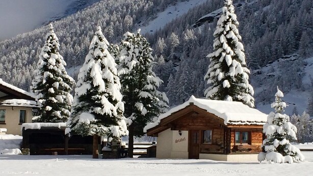 A snow-covered lodge with a wooden exterior, surrounded by snow-laden trees and a mountainous backdrop. - Chalet Stadel: Alpine Comfort (Saas Grund)