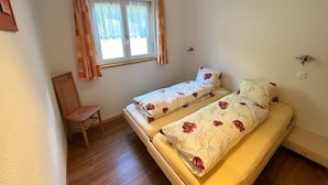 Une petite chambre avec un lit simple, un plancher en bois, une fenêtre avec des rideaux, une chaise et un radiateur fixé au mur.