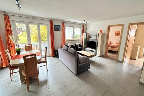 A modern living room with a sofa, a dining table, and chairs. There are large windows with curtains, a TV, and a bookshelf.