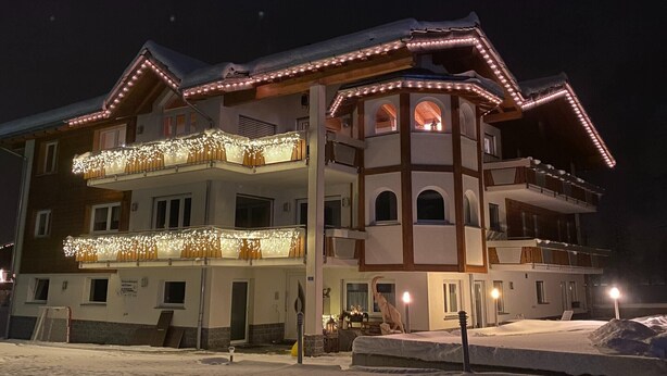 Un hôtel à plusieurs étages, doté de balcons décorés de guirlandes lumineuses, d’un toit enneigé et d’une tour centrale également pourvue d’un balcon.