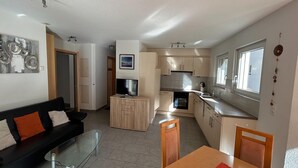 Een gezellig, compact leefruimte met een bank, een glazen salontafel, een televisie op een houten dressoir, een eettafel met stoelen, een keuken met kasten en een koelkast, en een raam.