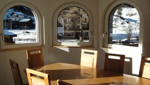 Een eethoek met een houten tafel en stoelen, drie gewelfde ramen met uitzicht op een besneeerd landschap en gebouwen.
