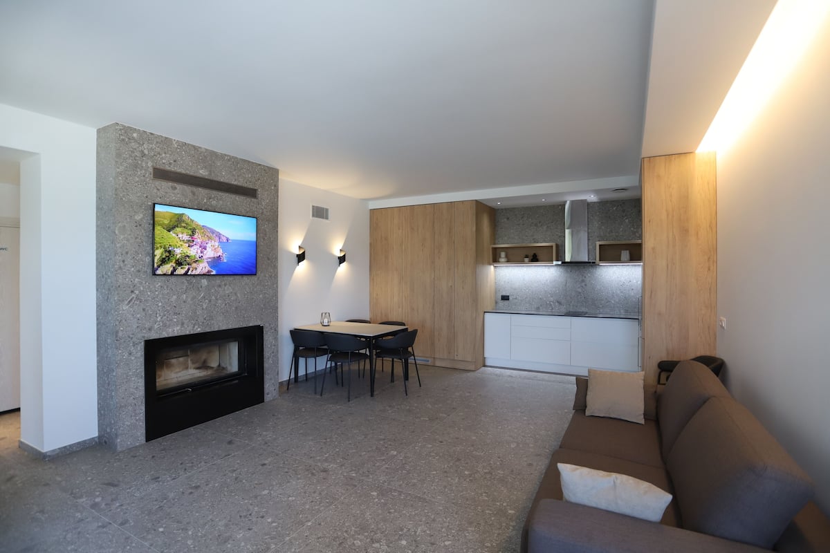 Una sala de estar moderna con chimenea, un televisor de pantalla plana montado en una pared de piedra, un comedor con mesa y sillas, y una cocina con armarios de madera y una encimera.