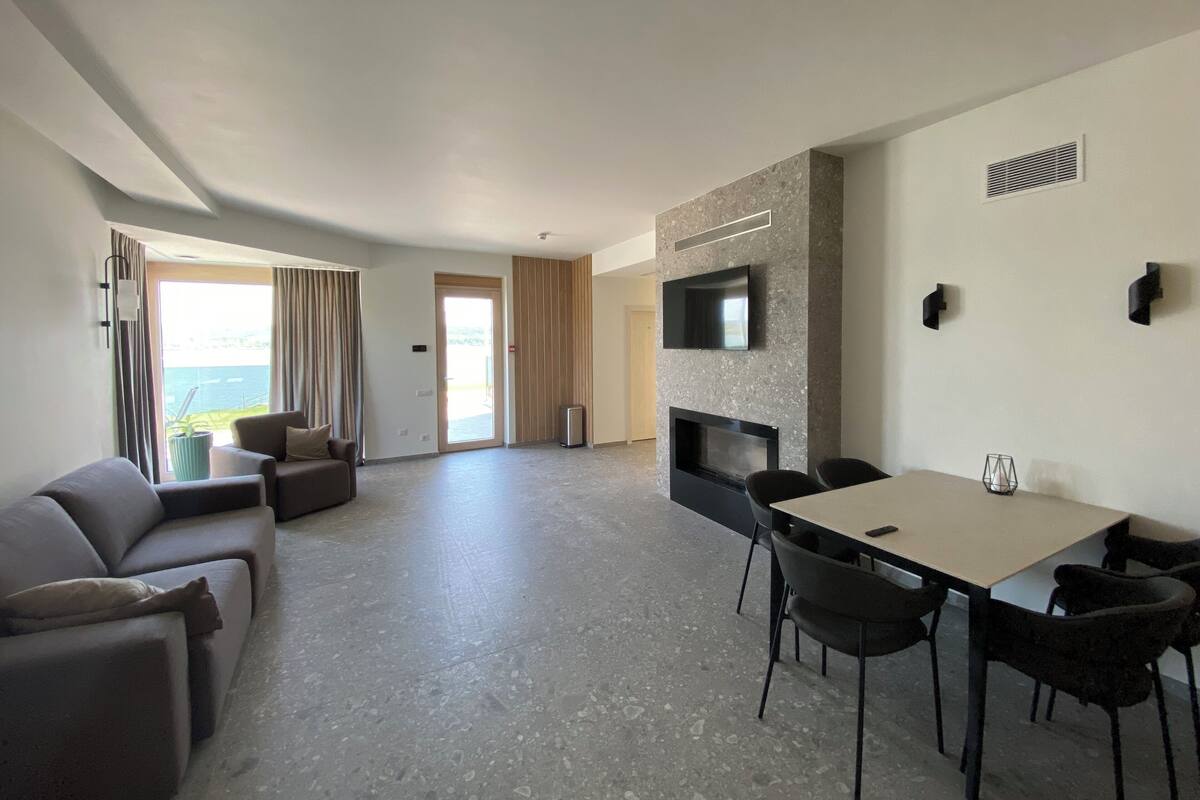 Una sala de estar moderna con un sofá, una mesa de comedor con sillas, un televisor de pantalla plana montado en una pared con textura y un puerta corrediza de vidrio que da a un balcón.