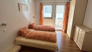 A small, single-room accommodation with a bed, a small cabinet, and a sliding glass door leading to a balcony.