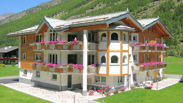 A multi-story hotel with balconies adorned with flowers, set against a mountainous backdrop.