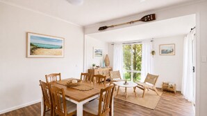 A dining area with a wooden table and chairs, a sideboard, and a view of the outdoors through a glass door. - Surfside - Beach Getaway (WOOLI)