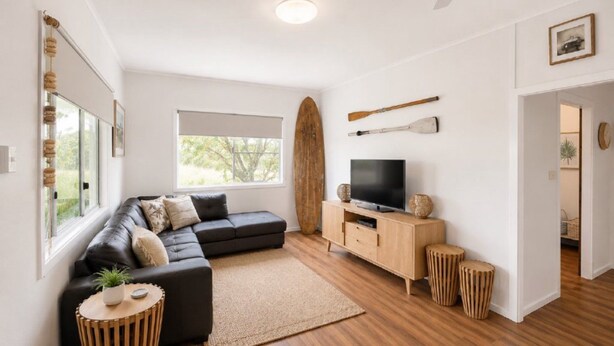 A modern living room with a sectional sofa, a flat-screen TV, wooden flooring, and a large window providing natural light. - Surfside - Beach Getaway (WOOLI)