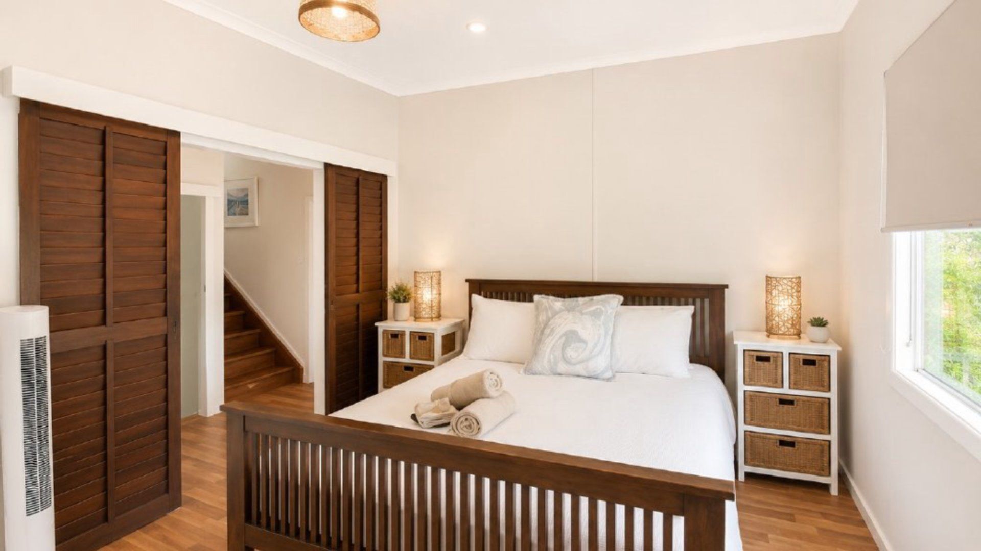 A bedroom with a wooden bed, white bedding, a nightstand with a lamp, and a window with a view.