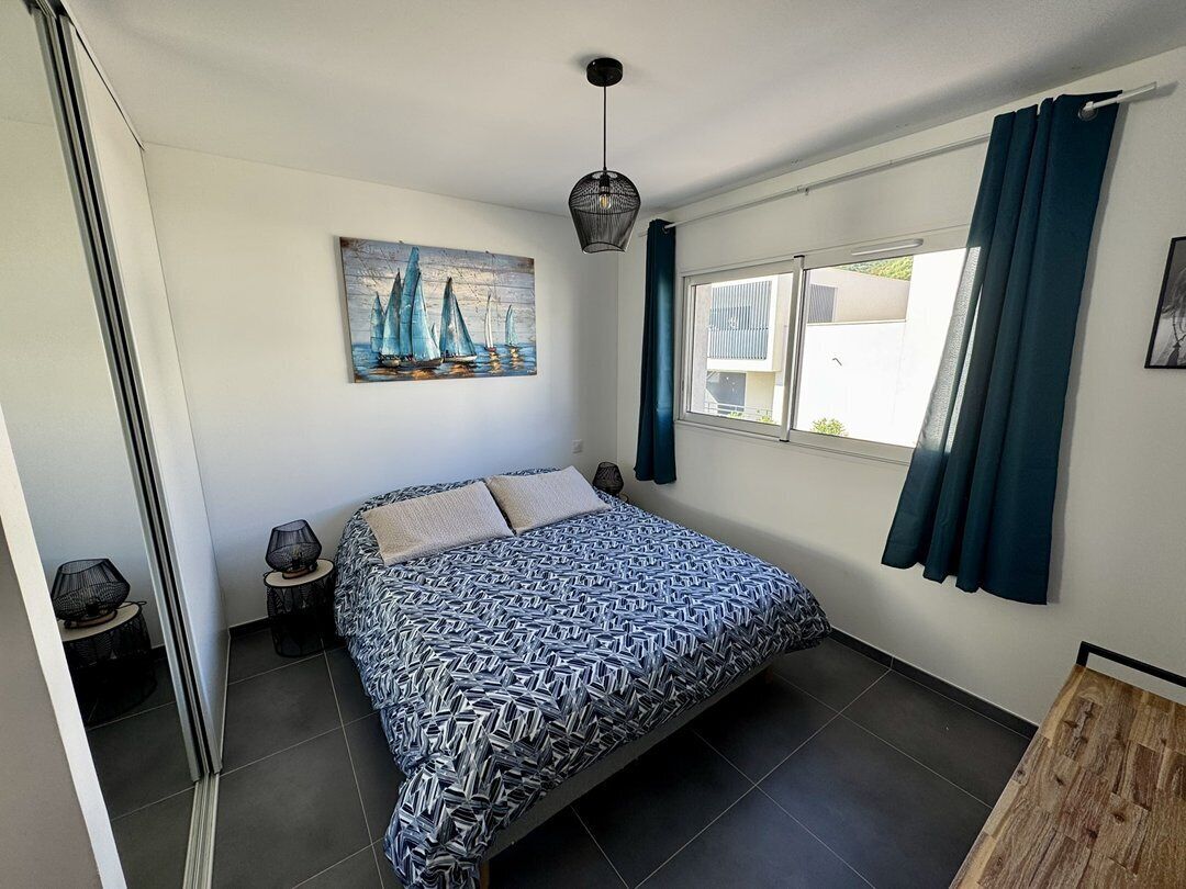 Un dormitorio con una cama, un cuadro, una ventana con cortinas y una mesita de noche con lámpara.