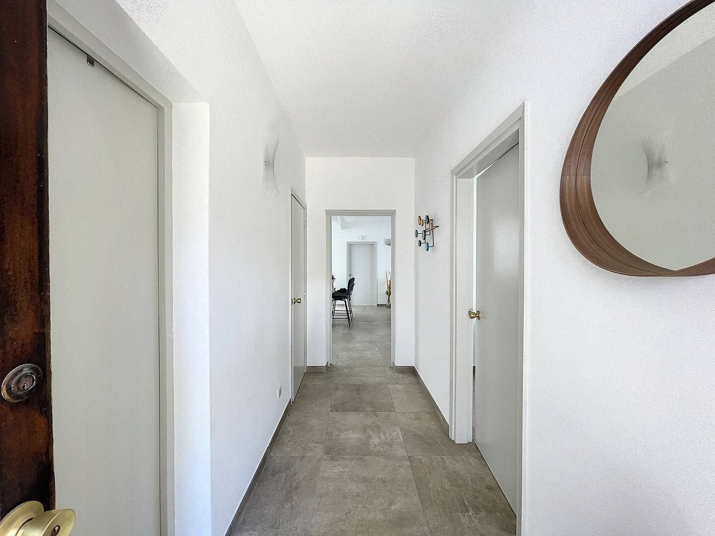 A hallway with white walls, a wooden door on the left, and a round mirror on the right.