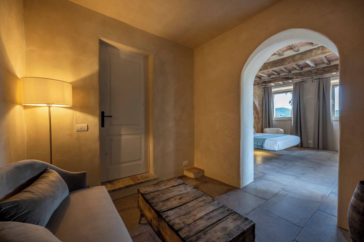 A cozy room with a sofa, a lamp, a wooden table, and an arched doorway leading to another room with a bed.