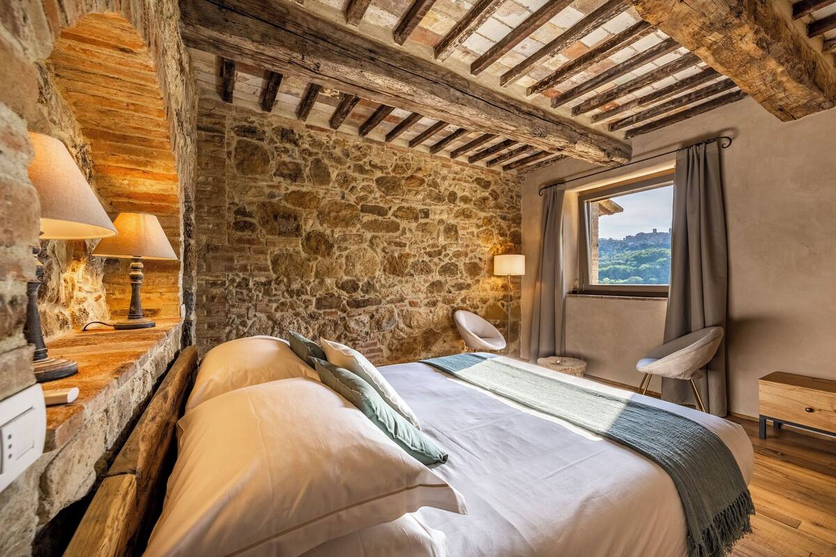 A bedroom with a stone wall, wooden beams, a bed with pillows, a bedside table with lamps, a chair, and a window with a view.