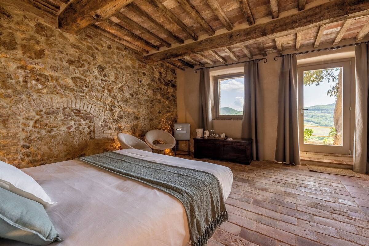 A rustic bedroom with a stone wall, wooden beams, a bed with a green throw, a chair, and a view of the countryside through a window.