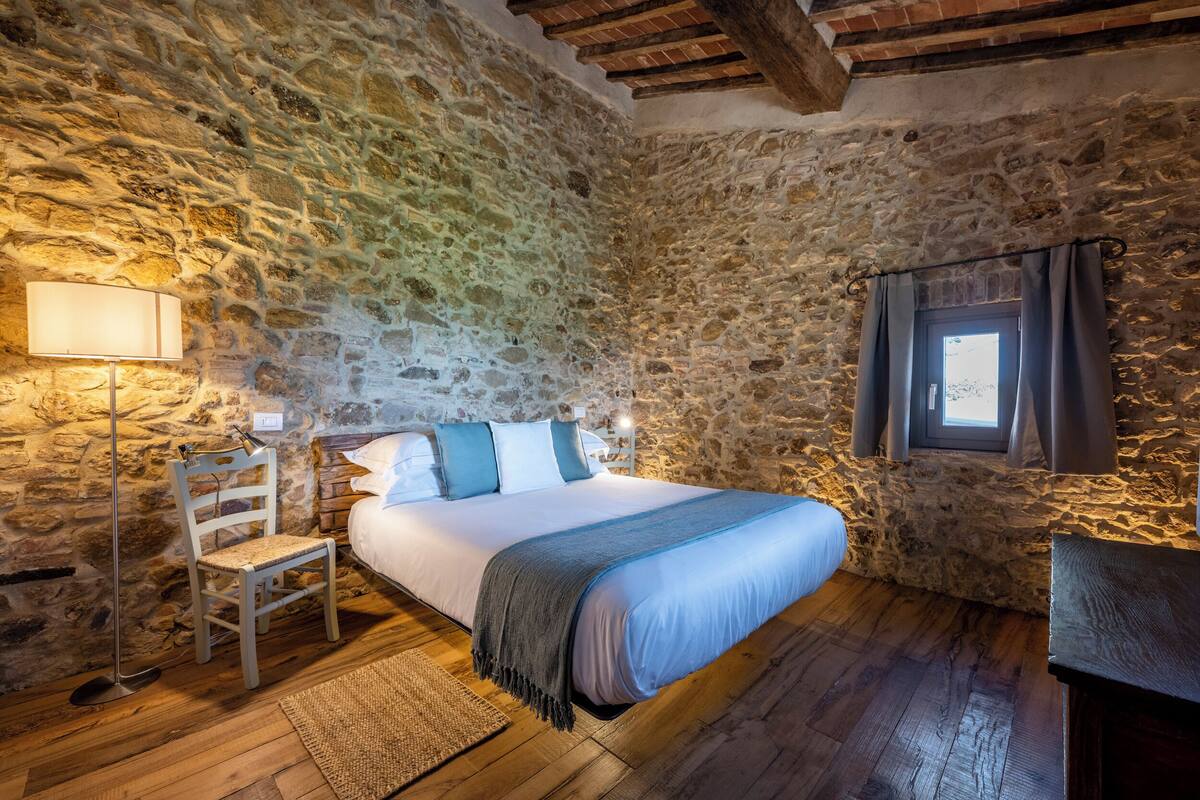 A bedroom with a stone wall, a wooden floor, a bed with a blue blanket, a chair, a lamp, and a window with curtains.