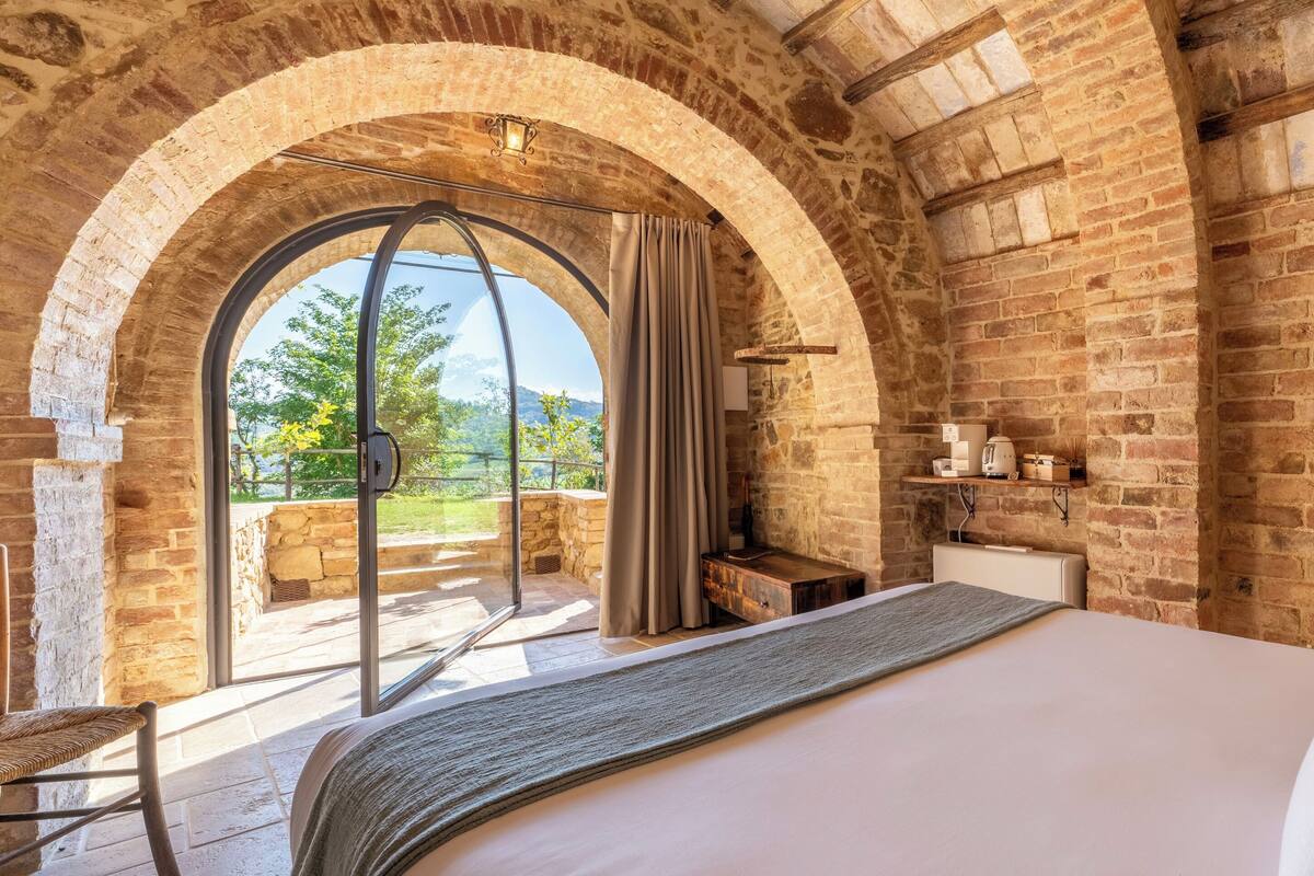 A bedroom with a large bed, a window with curtains, a wooden bench, and a brick wall.
