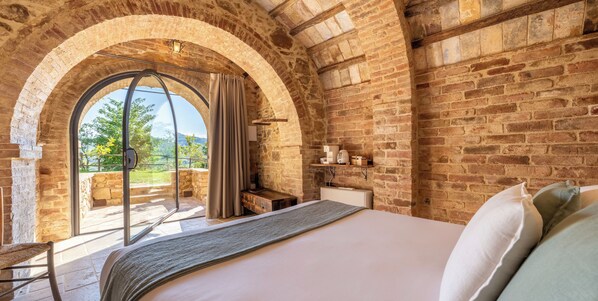 A bedroom with a large bed, a window with curtains, a wooden bench, and a brick wall. - Stelle di Monte (Montepulciano)